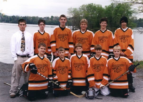 Ten players in orange and black jerseys and their coach pose by a lake, five kneeling in front.