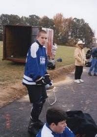 A young player in a blue jersey and pads stands on fresh asphalt, with grass and an open trailer behind.